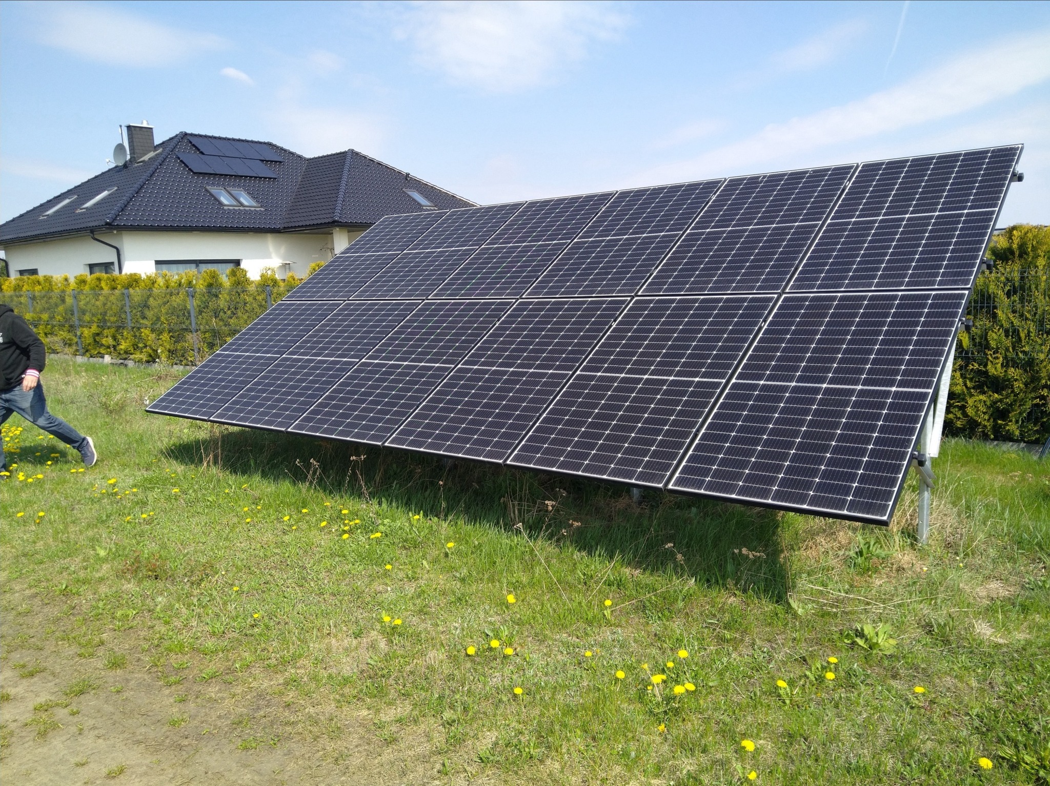 Panele fotowoltaiczne na trawniku przed domem z panelami na dachu, w tle osoba w bluzie przechodzi obok.
