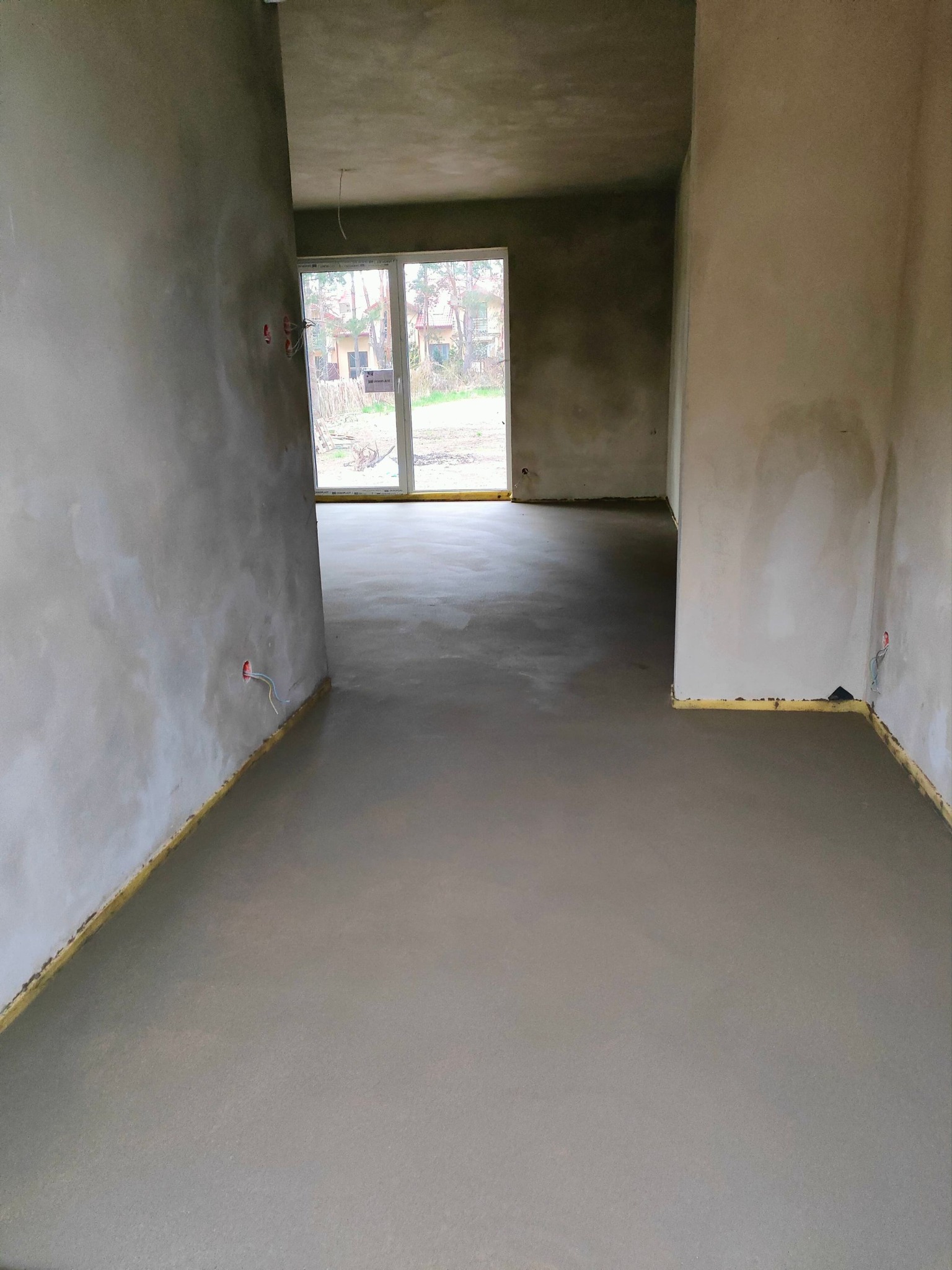 Świeżo wylana, gładka podłoga w surowym wnętrzu z widocznymi wystającymi przewodami elektrycznymi i piankową izolacją przy ścianach, z oknem balkonowym w tle.