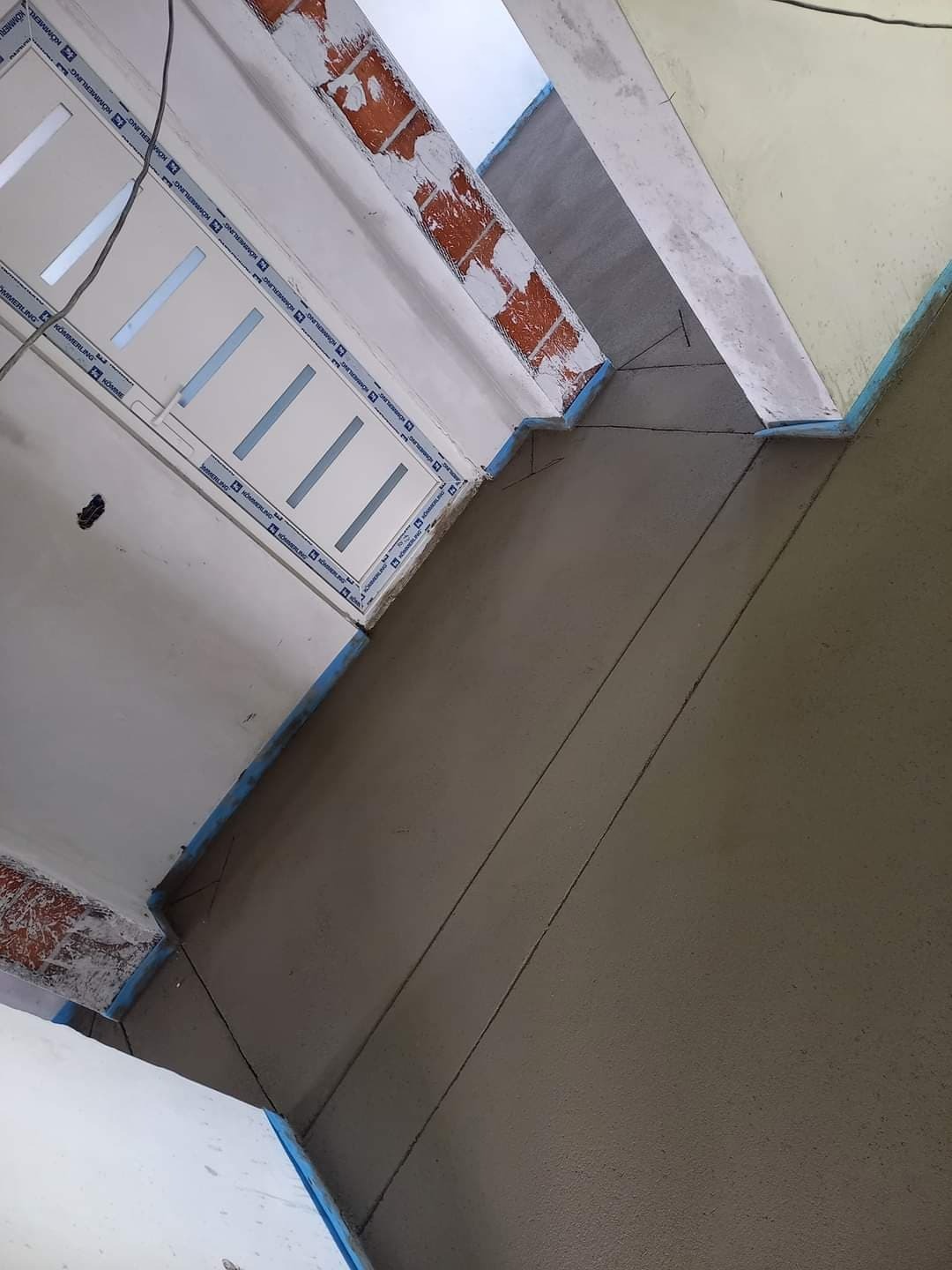 Świeżo wylana, gładka wylewka w pomieszczeniu z oknem, zabezpieczona niebieską taśmą malarską przy ścianach. Widoczne ślady po poziomowaniu i fragmenty cegieł na ścianie.