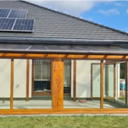 Zadaszony taras z drewnianą konstrukcją i przesuwnymi szklanymi panelami, widok z zewnątrz na trawnik i ogr&oacute;d, na dachu domu panele słoneczne.