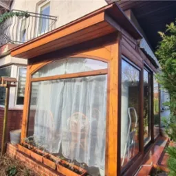 Drewniana zabudowa balkonu z przeszklonymi ścianami i firankami, widok z zewnątrz na tle ogrodu i elewacji domu.