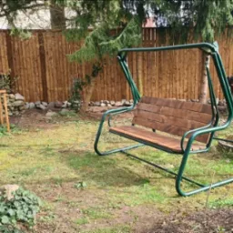 Metalowa huśtawka ogrodowa z drewnianym siedziskiem na tle drewnianego ogrodzenia i zielonego trawnika z widocznymi chwastami.