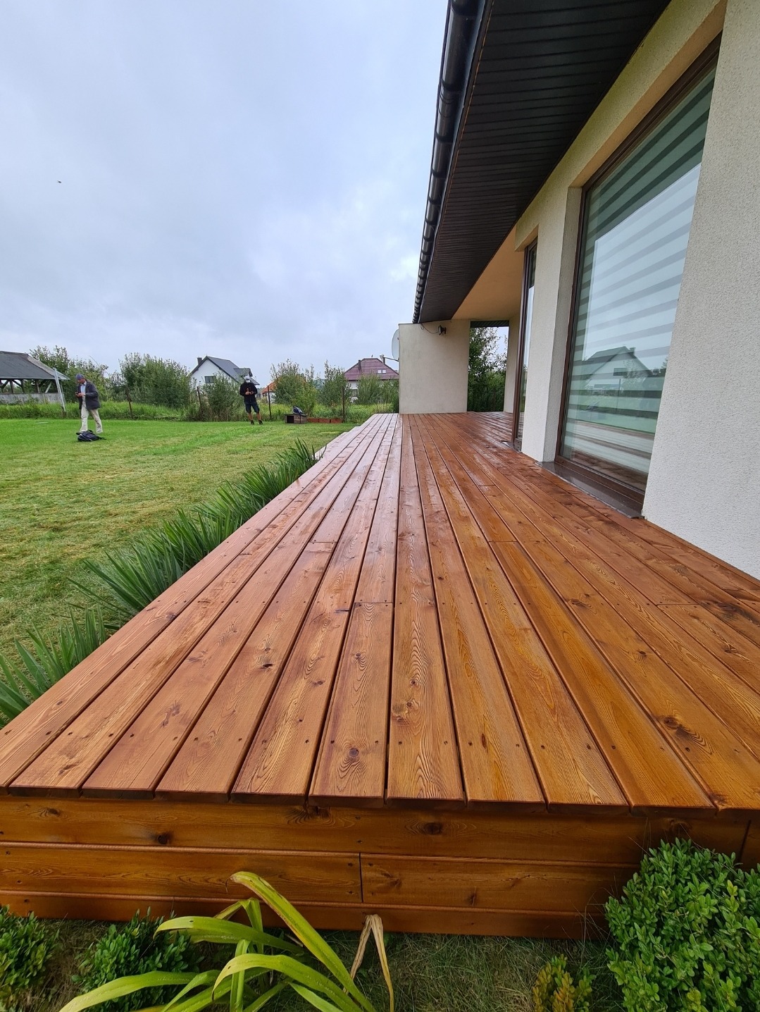 Świeżo zaimpregnowany drewniany taras przydomowy z widocznym usłojeniem desek, otoczony zadbanym trawnikiem i zielenią, w tle postacie dwóch osób na trawniku i domy jednorodzinne pod pochmurnym...