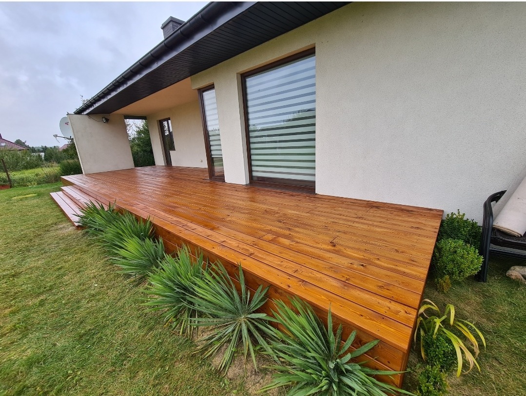 Drewniany taras przy domu, wykończony na wysoki połysk, z niskimi schodkami i roślinami ozdobnymi wzdłuż krawędzi.