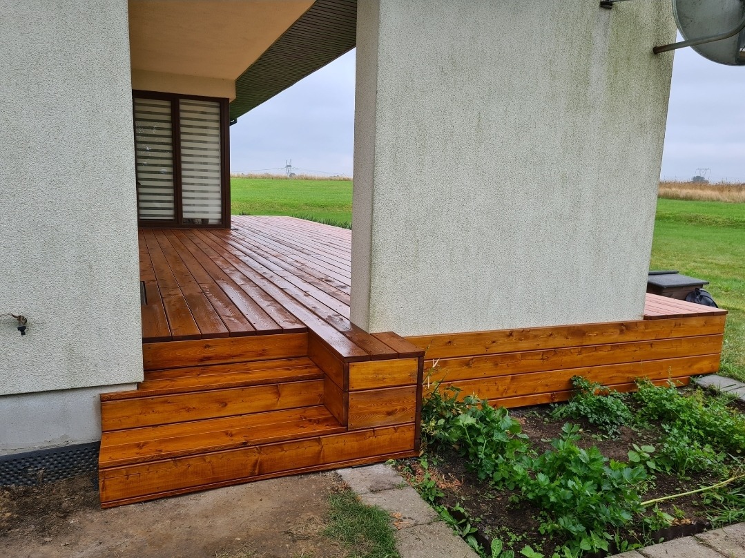 Świeżo zaimpregnowany taras drewniany z trzema stopniami prowadzącymi na podest, widoczne krople wody na deskach, obok domu z szarą elewacją i zielenią wokół.