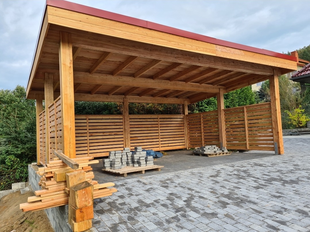 Drewniana wiata z ażurowymi ścianami i czerwonym dachem, w trakcie budowy, z paletami z kostką brukową i stosem drewna na pierwszym planie.