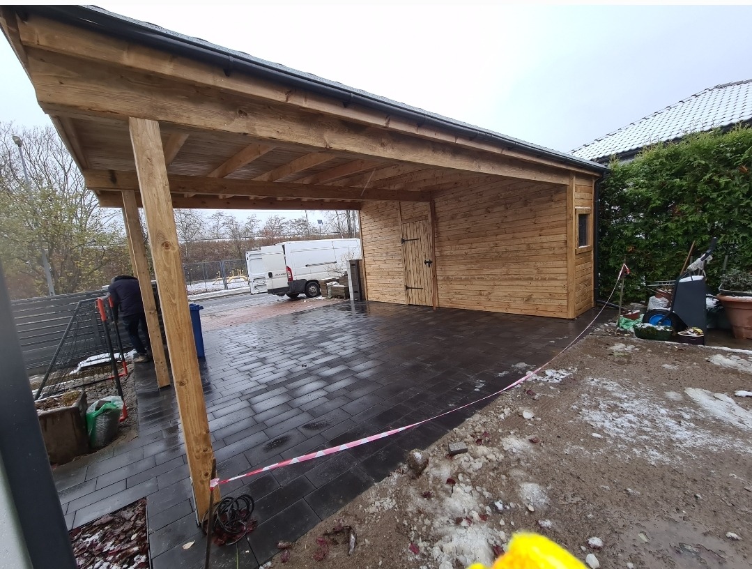 Drewniana wiata garażowa z pomieszczeniem gospodarczym, wykończona deską boazeryjną, z czarną dachówką i podjazdem z ciemnej kostki brukowej, w tle biały samochód dostawczy i częściowo ośnieżony...