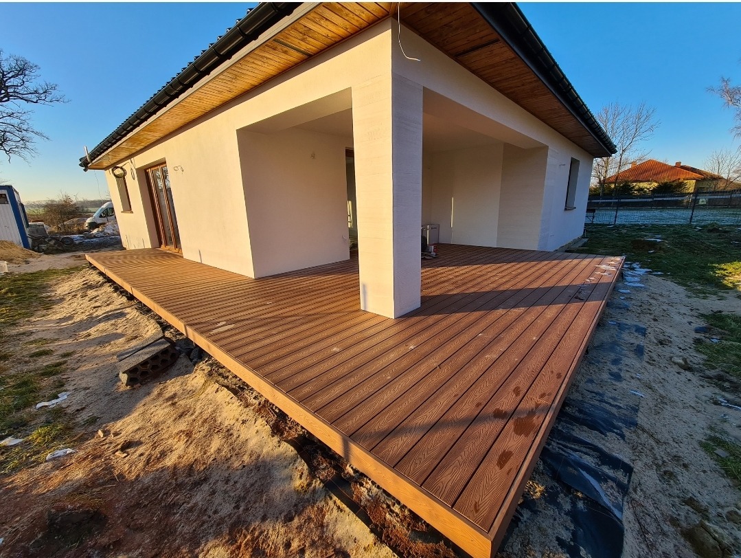 Rozległy, brązowy taras kompozytowy przylegający do jasnego domu z kolumnami, widoczny w zimowym świetle, z fragmentami śniegu na trawie w tle.