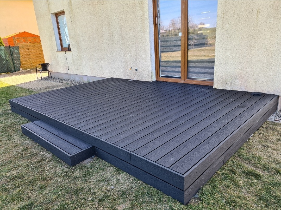 Nowoczesny, grafitowy taras z desek kompozytowych z dwustopniowymi schodkami, przylegający do jasnej ściany budynku z oknem i przeszklonymi drzwiami tarasowymi, otoczony trawnikiem.