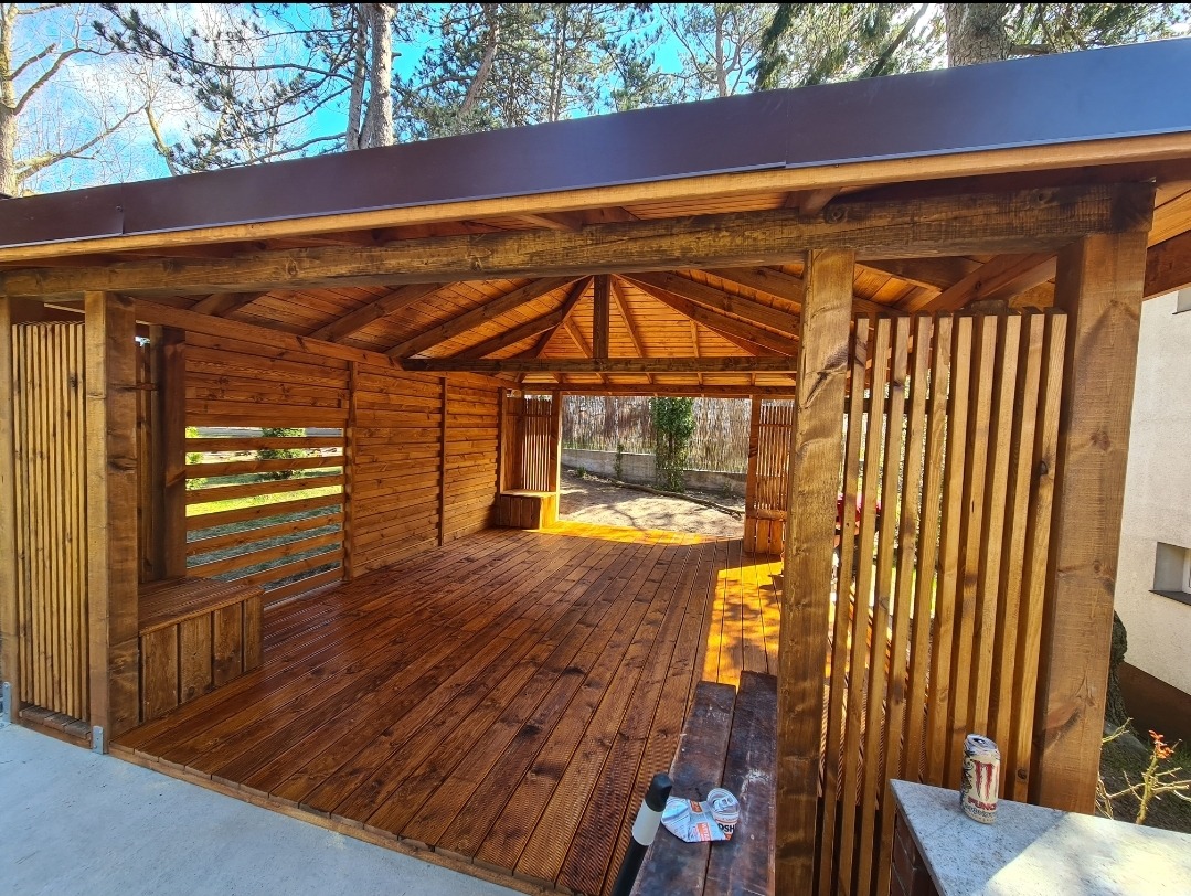 Drewniana altana ogrodowa z brązowym zadaszeniem, widoczne elementy konstrukcyjne, podłoga i ściany z desek, na podłodze puszki po napojach, w tle zieleń ogrodu.