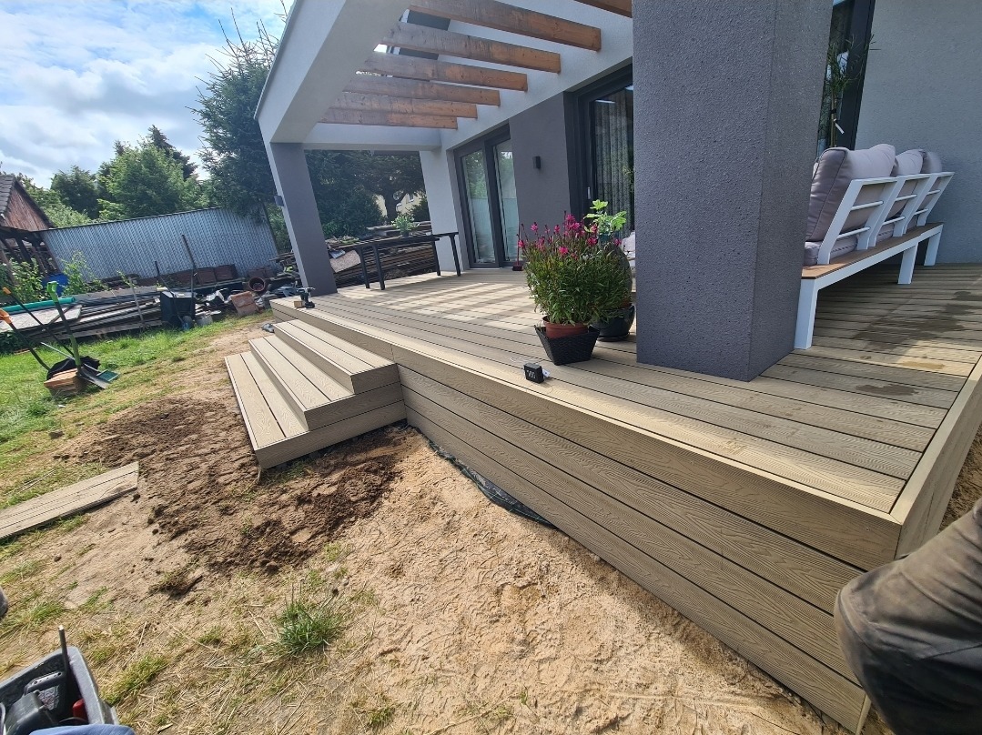 Nowo wybudowany taras z desek kompozytowych z niskimi schodami i pergolą, częściowo zagospodarowany teren wokół, widoczne ślady prac budowlanych.