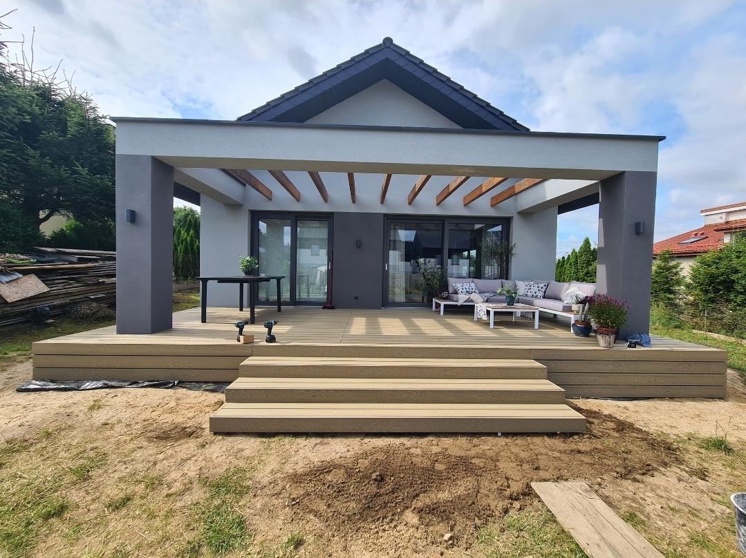 Nowoczesny taras drewniany z kompozytu zadaszony pergolą, z meblami ogrodowymi i schodami, w trakcie montażu widoczne wkrętarki.