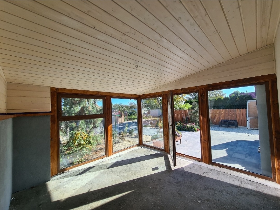 Wnętrze przeszklonego balkonu lub tarasu z drewnianą konstrukcją, widok na ogród i patio z kostki brukowej, surowy stan podłogi.