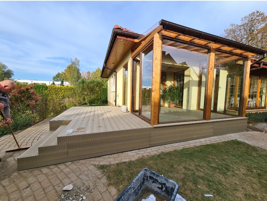 Jasny, kompozytowy taras przylegający do domu z przeszkloną werandą; mężczyzna zamiata powierzchnię tarasu, widoczny także fragment muru i trawnika.