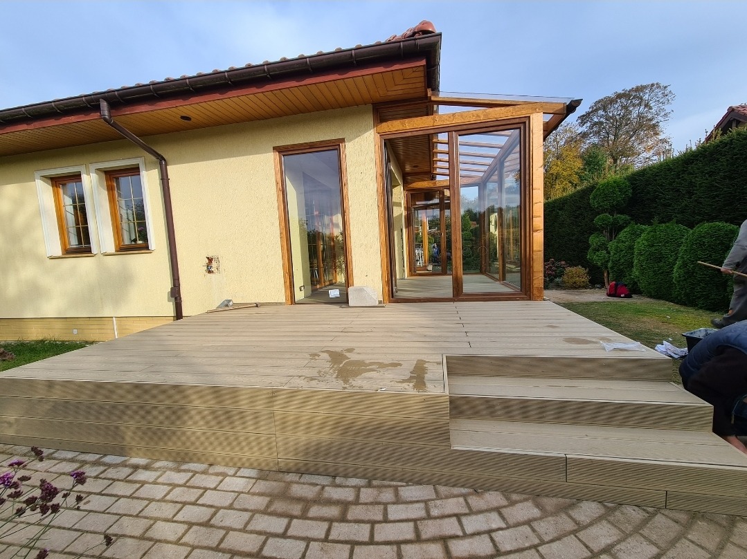 Jasny taras z kompozytu z dwustopniowymi schodami, przylegający do domu z przeszklonym ogrodem zimowym z drewnianą konstrukcją, widok na kostkę brukową i ogród.