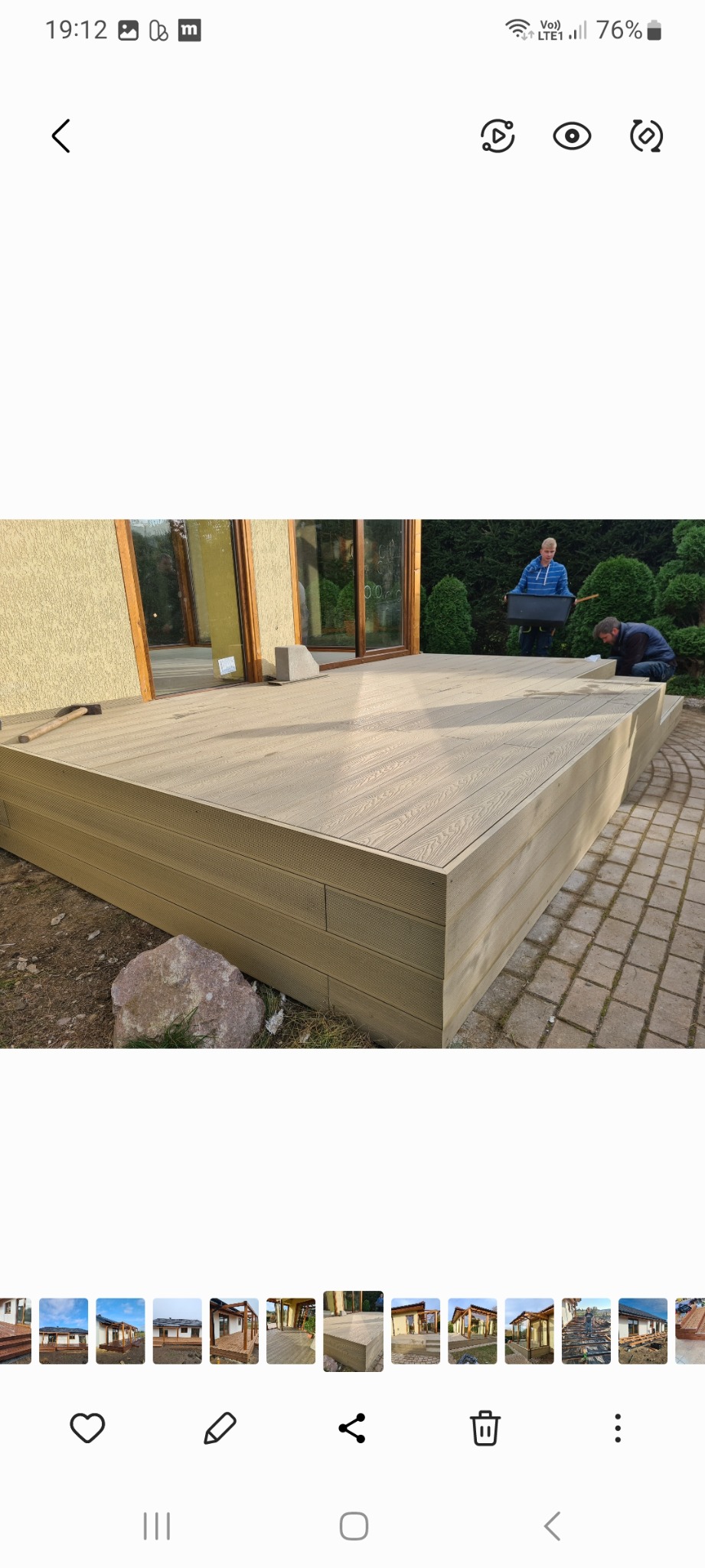 Budowa tarasu z kompozytu drewnopodobnego: dwóch mężczyzn pracuje nad montażem desek na przygotowanej konstrukcji, widoczny młotek i poziomica, w tle budynek z dużymi oknami i zadbana roślinność.