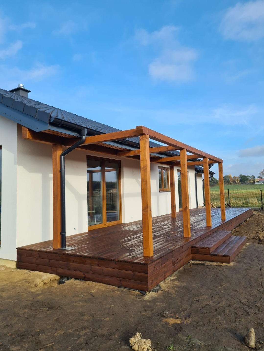 Nowoczesny taras z ciemnego drewna z zadaszeniem, przylegający do białego domu z czarnym dachem, widok na ogród w tle.