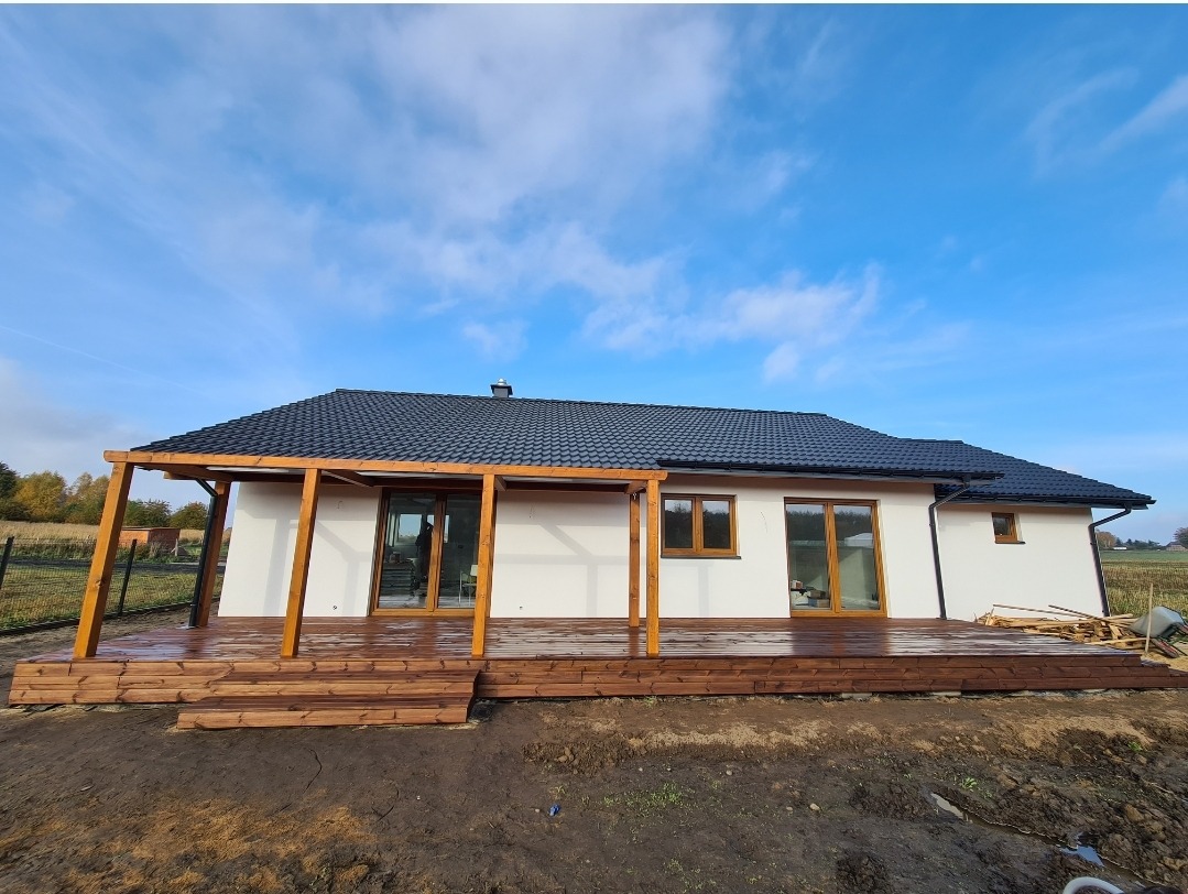 Nowoczesny dom jednorodzinny z drewnianym tarasem i pergolą z belek o przekroju kwadratowym, widok z poziomu gruntu na tle błękitnego nieba z chmurami.