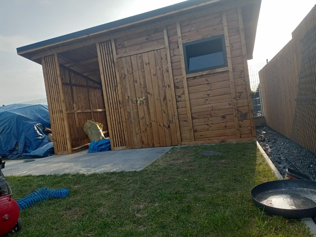 Brązowa drewniana szopa ogrodowa z drzwiami i małym oknem, częściowo otwarta z boku, stojąca na trawniku obok drewnianego ogrodzenia, z widocznymi narzędziami i workiem w środku.