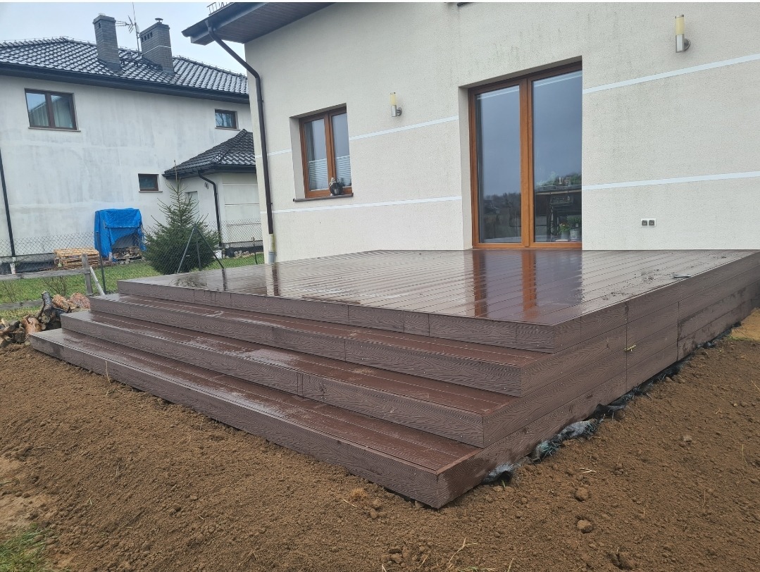 Nowo wybudowany, brązowy taras z desek kompozytowych, prowadzący do drzwi balkonowych w jasnym domu. Widoczne trzy stopnie i mokra nawierzchnia tarasu.