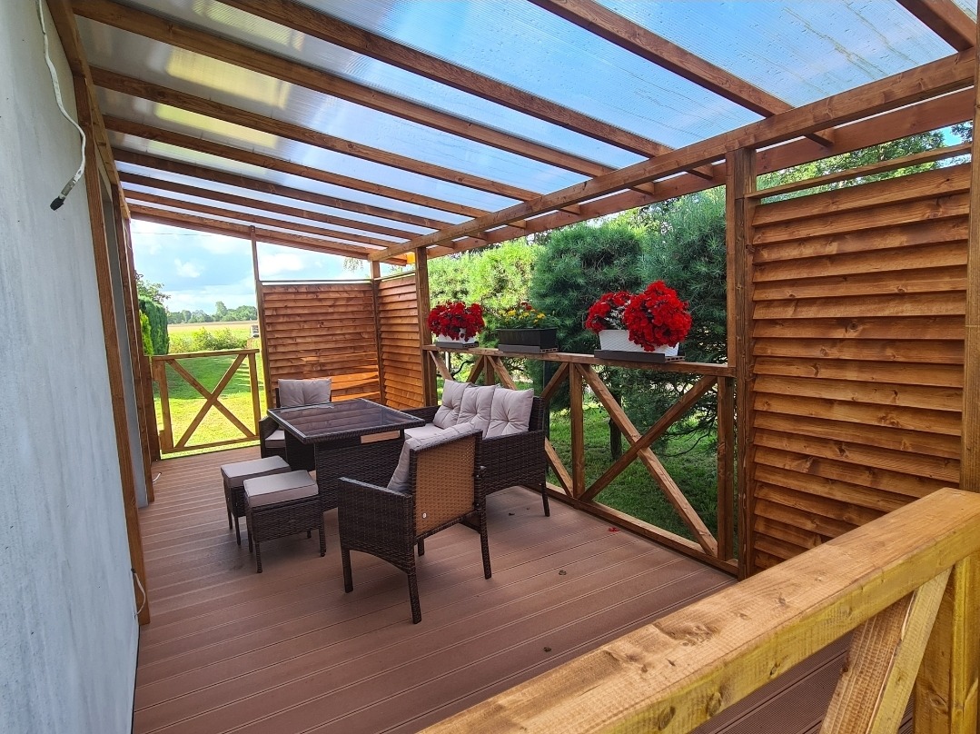 Zadaszony taras drewniany z meblami ogrodowymi w Koszalinie, z widokiem na zieleń, dekorowany czerwonymi kwiatami w donicach.