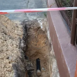 Wąski wykop w ziemi, zabezpieczony taśmą ostrzegawczą, z widoczną rurą instalacyjną na dnie, tuż obok ogrodzenia posesji.