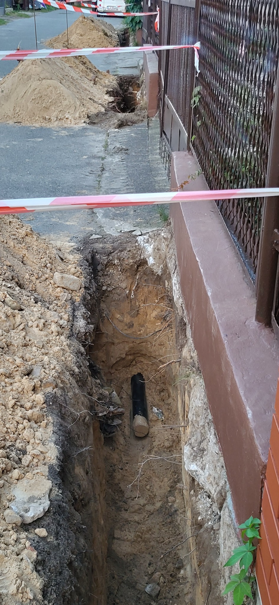 Wąski wykop w ziemi, zabezpieczony taśmą ostrzegawczą, z widoczną rurą instalacyjną na dnie, tuż obok ogrodzenia posesji.