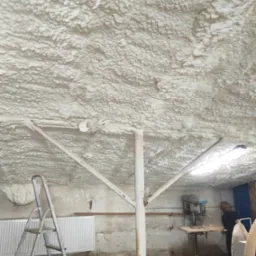 Strop pokryty grubą warstwą izolacji, widoczne elementy konstrukcyjne i instalacja elektryczna, w tle warsztat z wiertarką stołową.