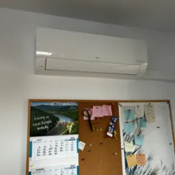 Biały klimatyzator LG Dual Inverter zamontowany na ścianie nad korkową tablicą z kalendarzami i notatkami w biurze.