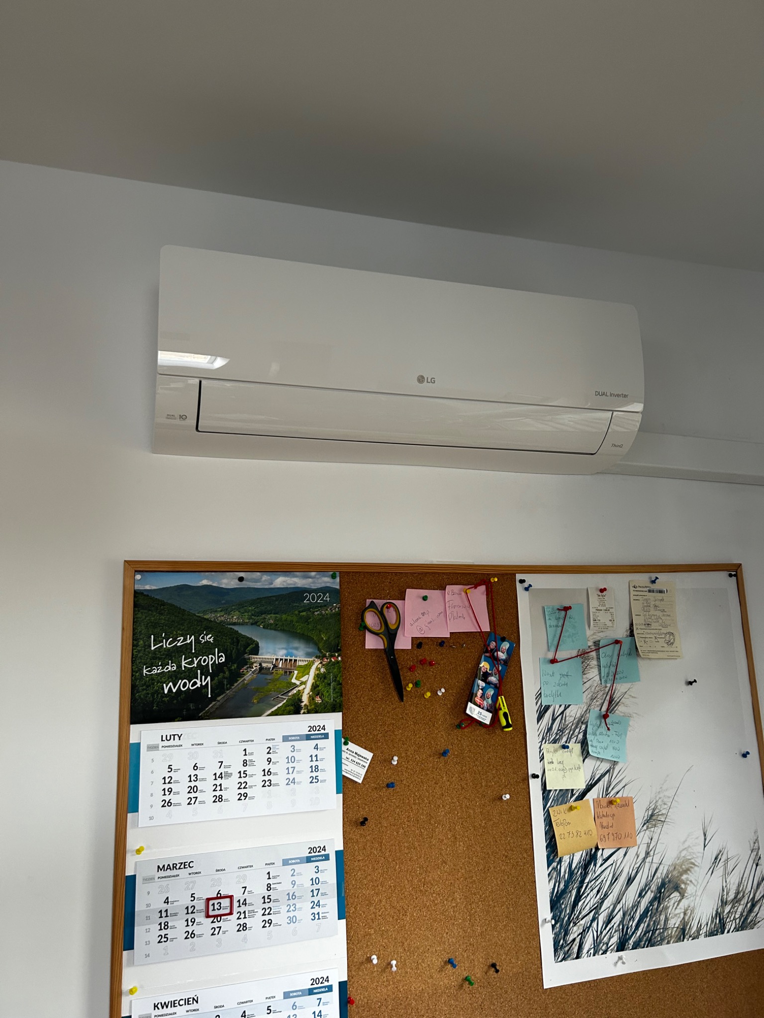 Biały klimatyzator LG typu split na ścianie w biurze, pod nim tablica korkowa z kalendarzem i notatkami.