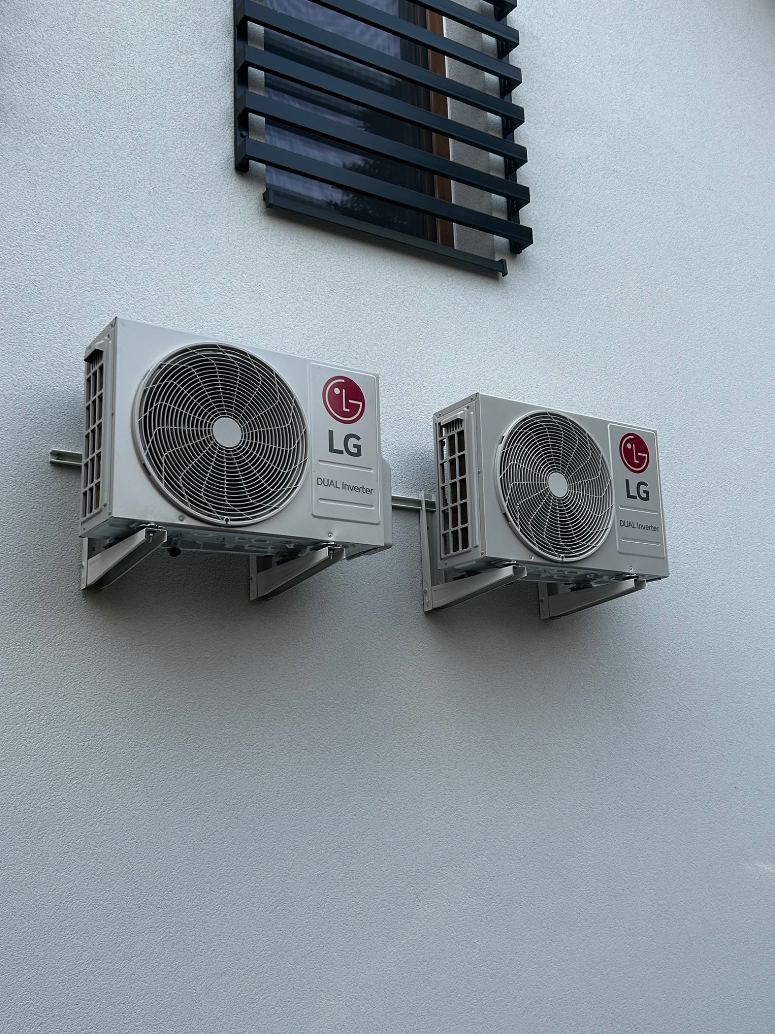 Dwa identyczne, białe klimatyzatory LG Dual Inverter zamontowane na zewnętrznej, szarej ścianie budynku z widocznym fragmentem czarnej, metalowej konstrukcji okiennej nad nimi.
