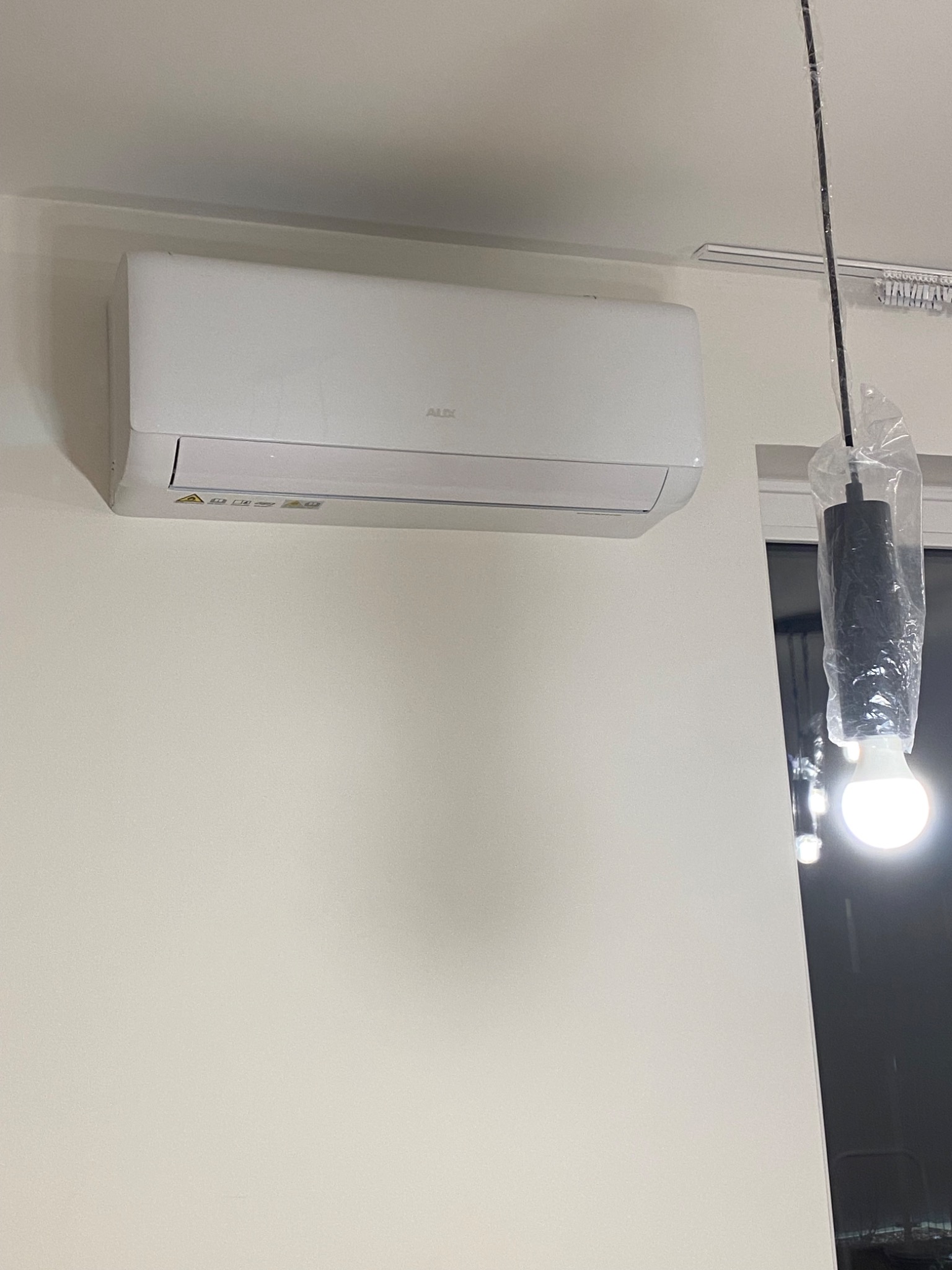 Biała jednostka klimatyzacji marki AUX zamontowana na ścianie w jasnym pomieszczeniu, widoczna lampa wisząca w folii ochronnej.