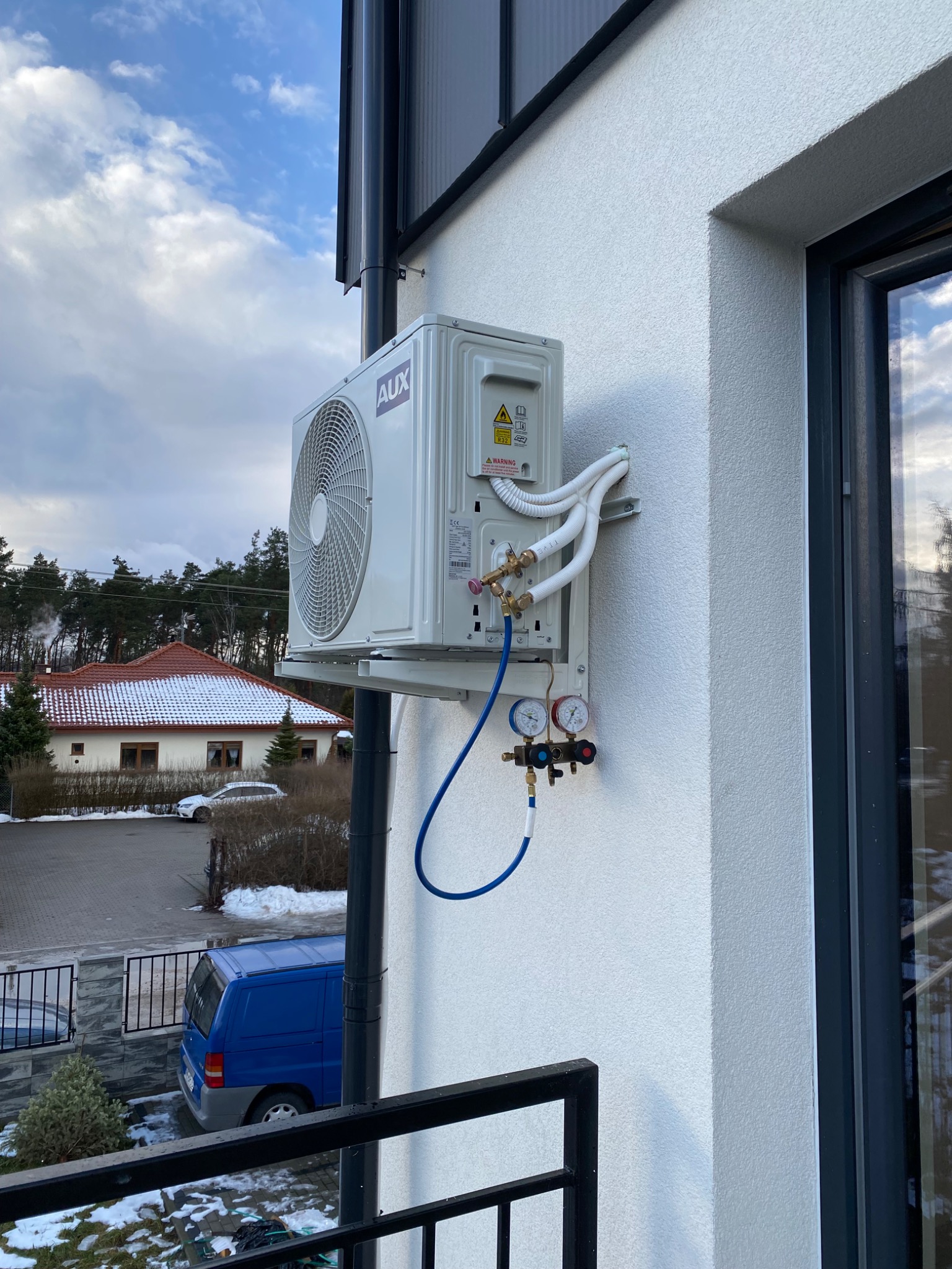 Biała jednostka zewnętrzna klimatyzacji AUX zamontowana na elewacji domu, podłączona do manometru serwisowego, w tle widoczny ośnieżony dach sąsiedniego budynku i błękitny samochód dostawczy.