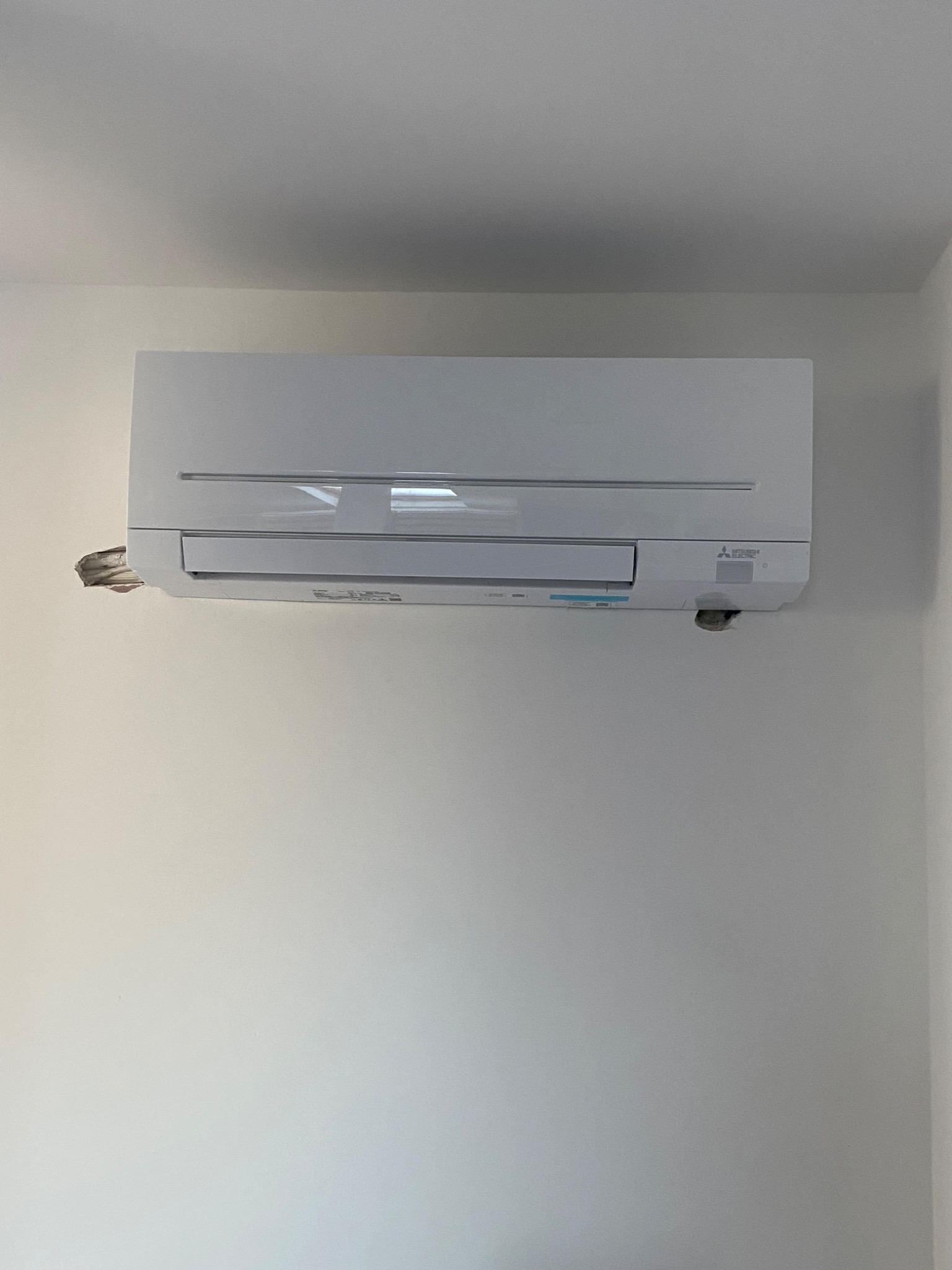 Biały klimatyzator Mitsubishi Electric zamontowany na białej ścianie, widoczne niedoskonałości w wykończeniu ściany wokół przewodów.
