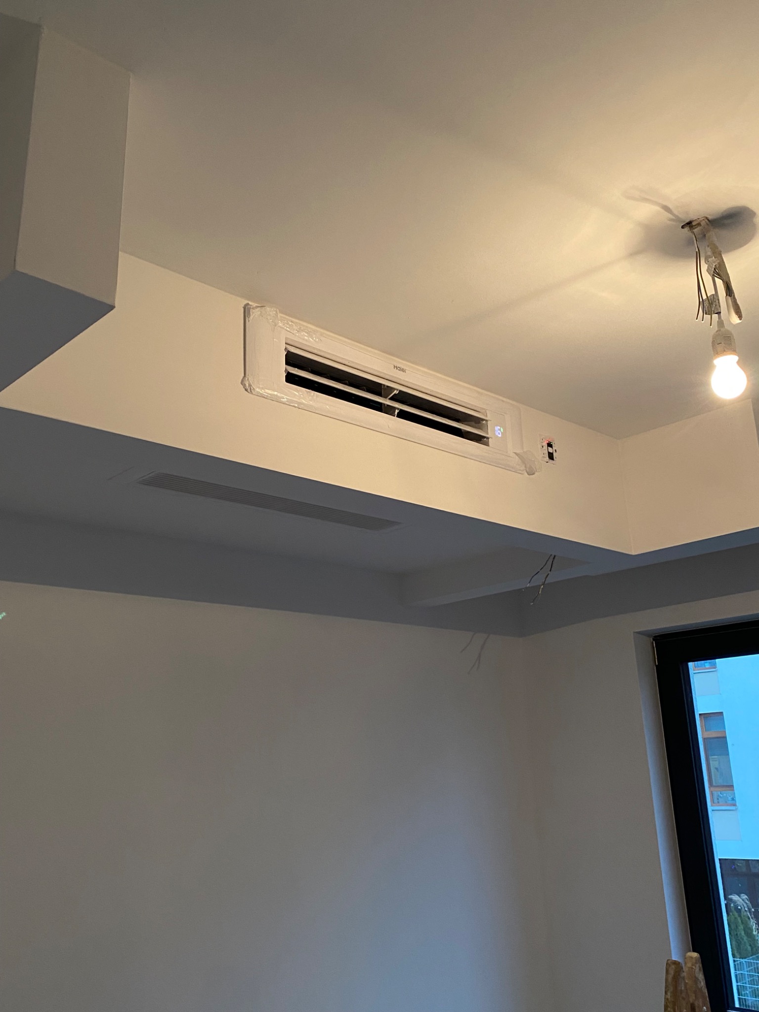 Biała klimatyzacja marki Haier zamontowana na suficie w pomieszczeniu w trakcie prac wykończeniowych, widoczne kable i elementy instalacji oświetleniowej oraz częściowo zabezpieczona folią ochronną.