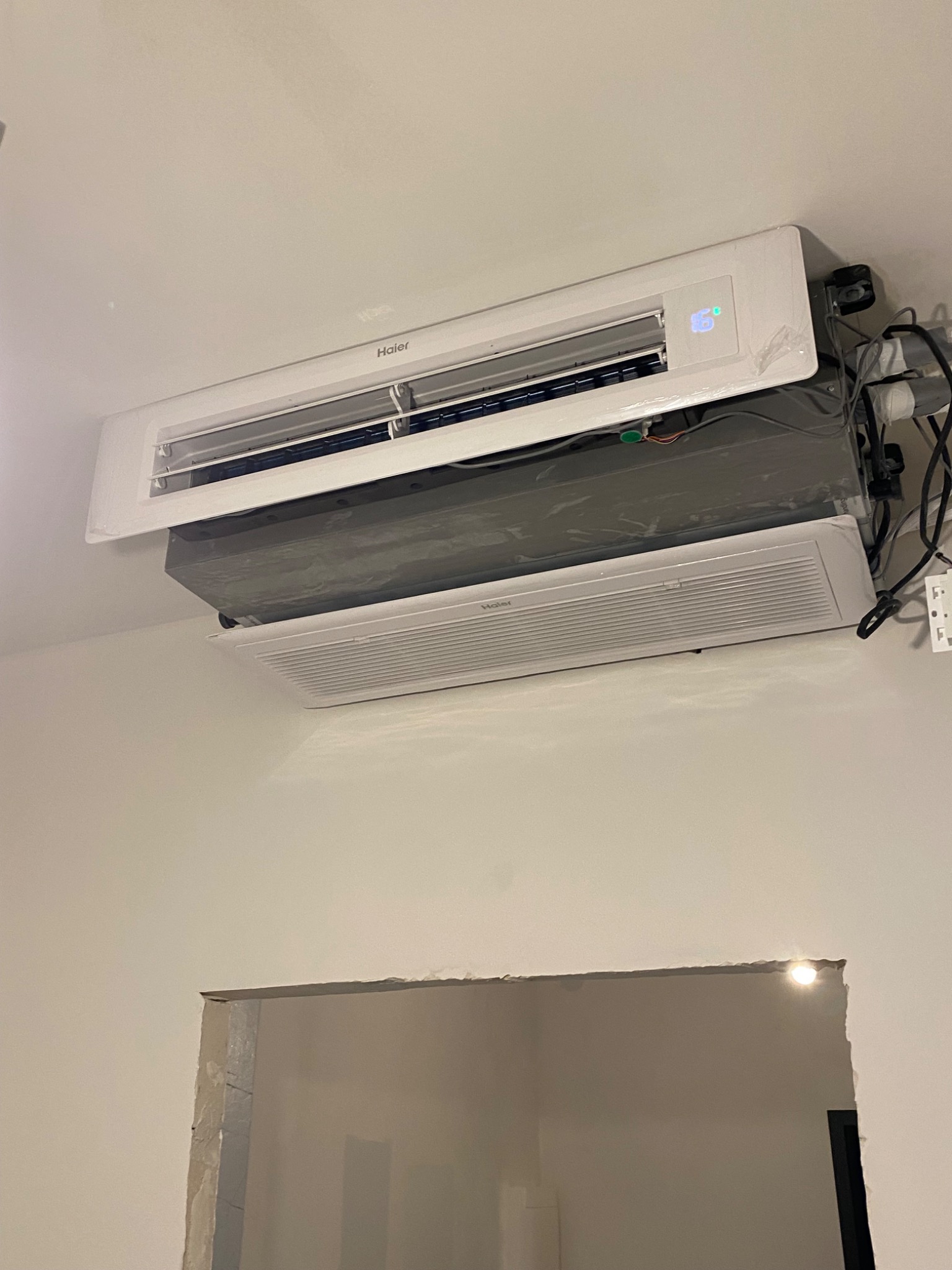Dwa klimatyzatory zamontowane pod sufitem, jeden marki Haier z wyświetlaczem 16 stopni, drugi pod nim, jeszcze w folii ochronnej, widoczne przewody i niedokończone wykończenie ściany z otworem.