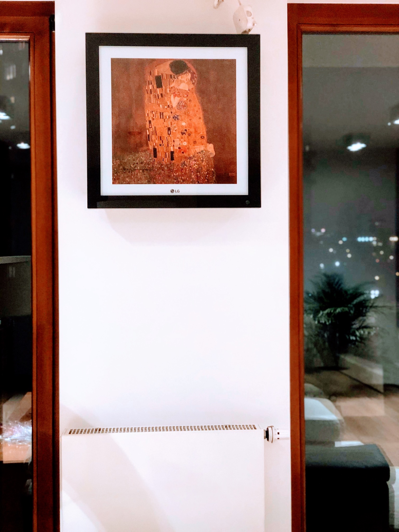 Telewizor LG The Frame wyświetlający obraz Gustava Klimta 'Pocałunek' w salonie z grzejnikiem i oknem na tle nocnego miasta.