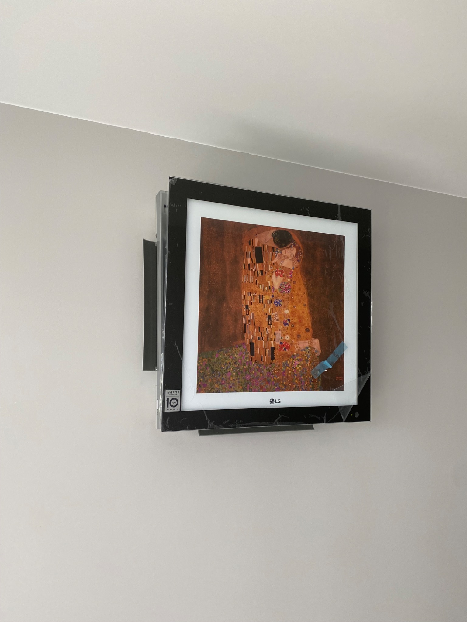 Telewizor LG z czarną ramką, wyświetlający obraz Klimta 'Pocałunek', zamontowany na ścianie, jeszcze w folii ochronnej.