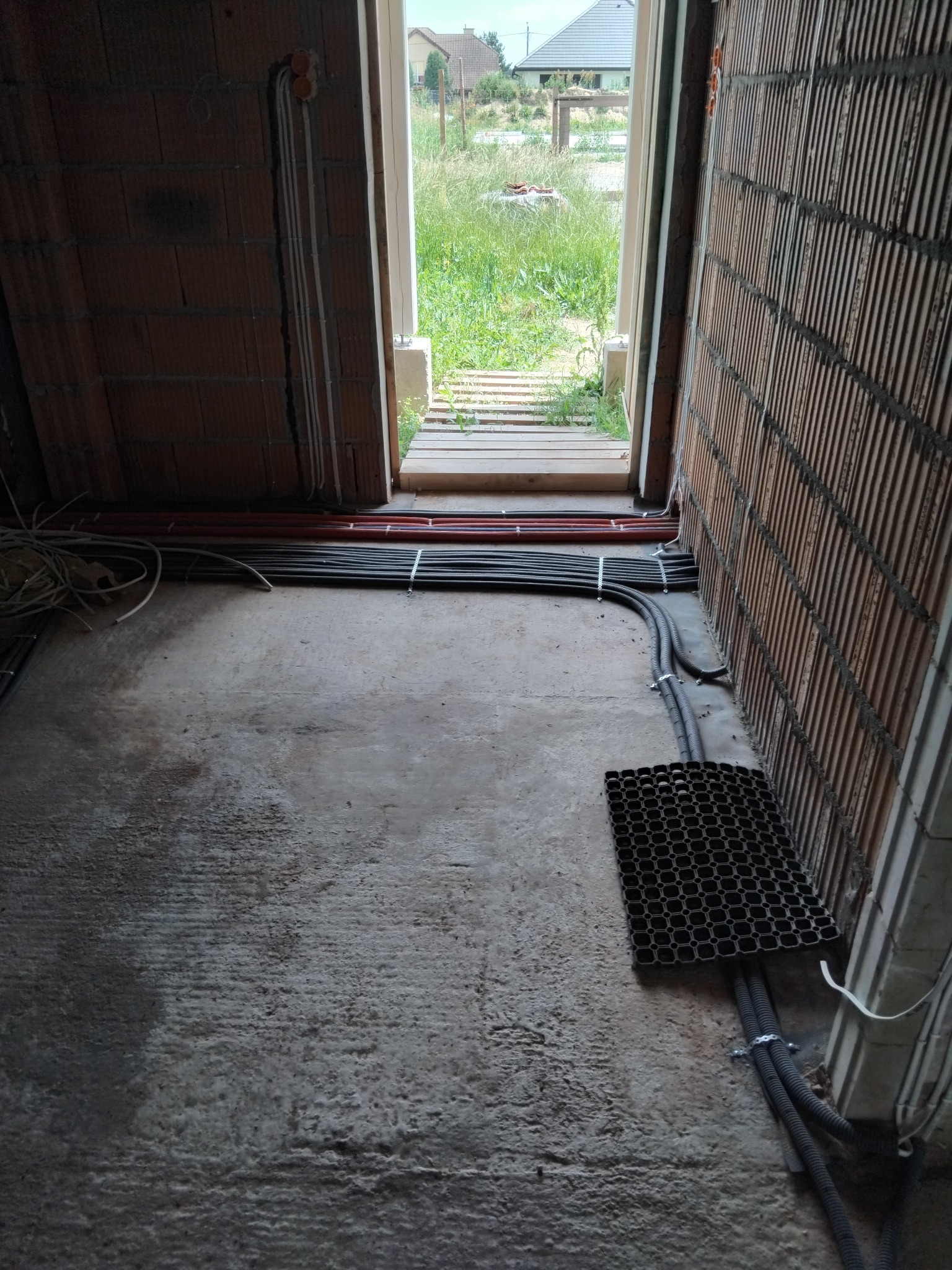 Wnętrze w stanie surowym z widocznymi przewodami elektrycznymi ułożonymi w korytkach i przy ścianach z cegły dziurawki, widok na zewnątrz przez otwarte drzwi.