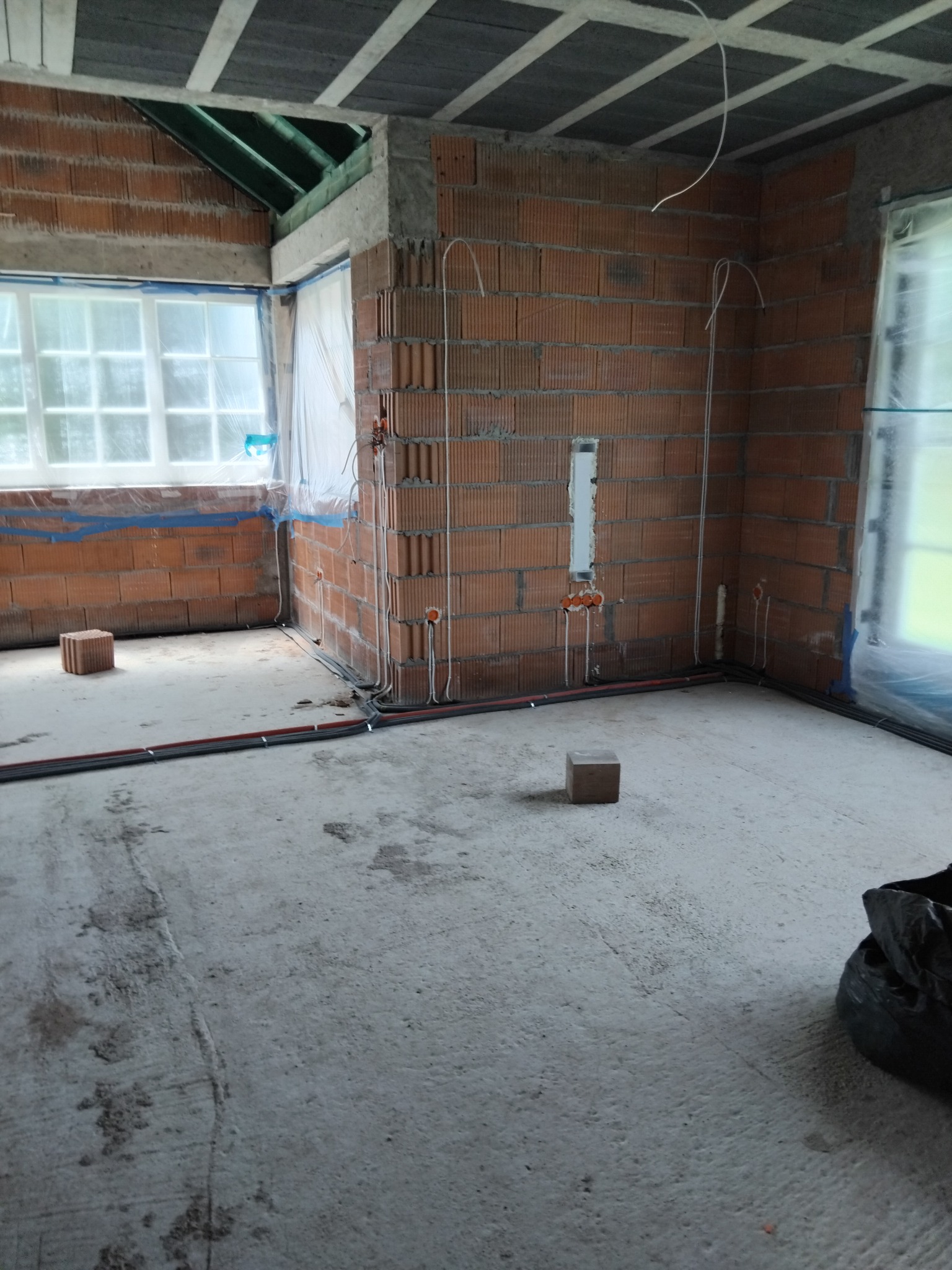 Wnętrze w stanie surowym: ściany z cegły, instalacja elektryczna w peszlach, okna zabezpieczone folią, betonowa podłoga z widocznymi śladami prac budowlanych.