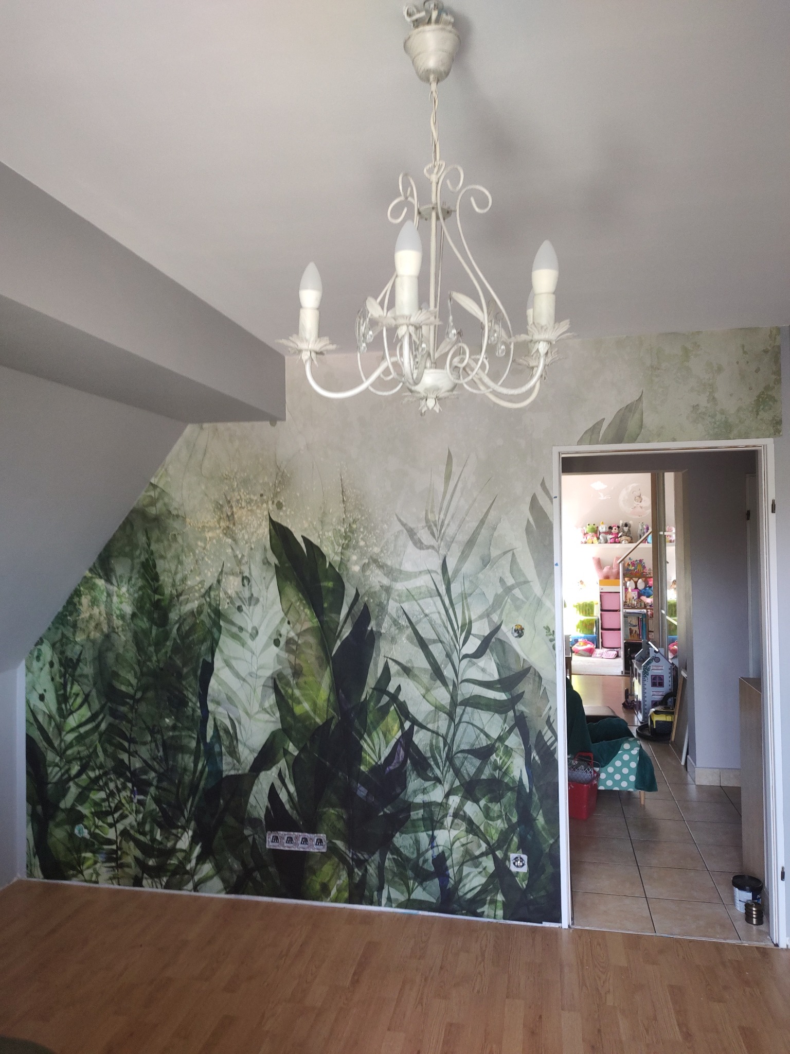 Pokój z fototapetą z motywem dżungli, kremowy żyrandol z kryształkami, drewniana podłoga i widok na pokój dziecięcy w tle.