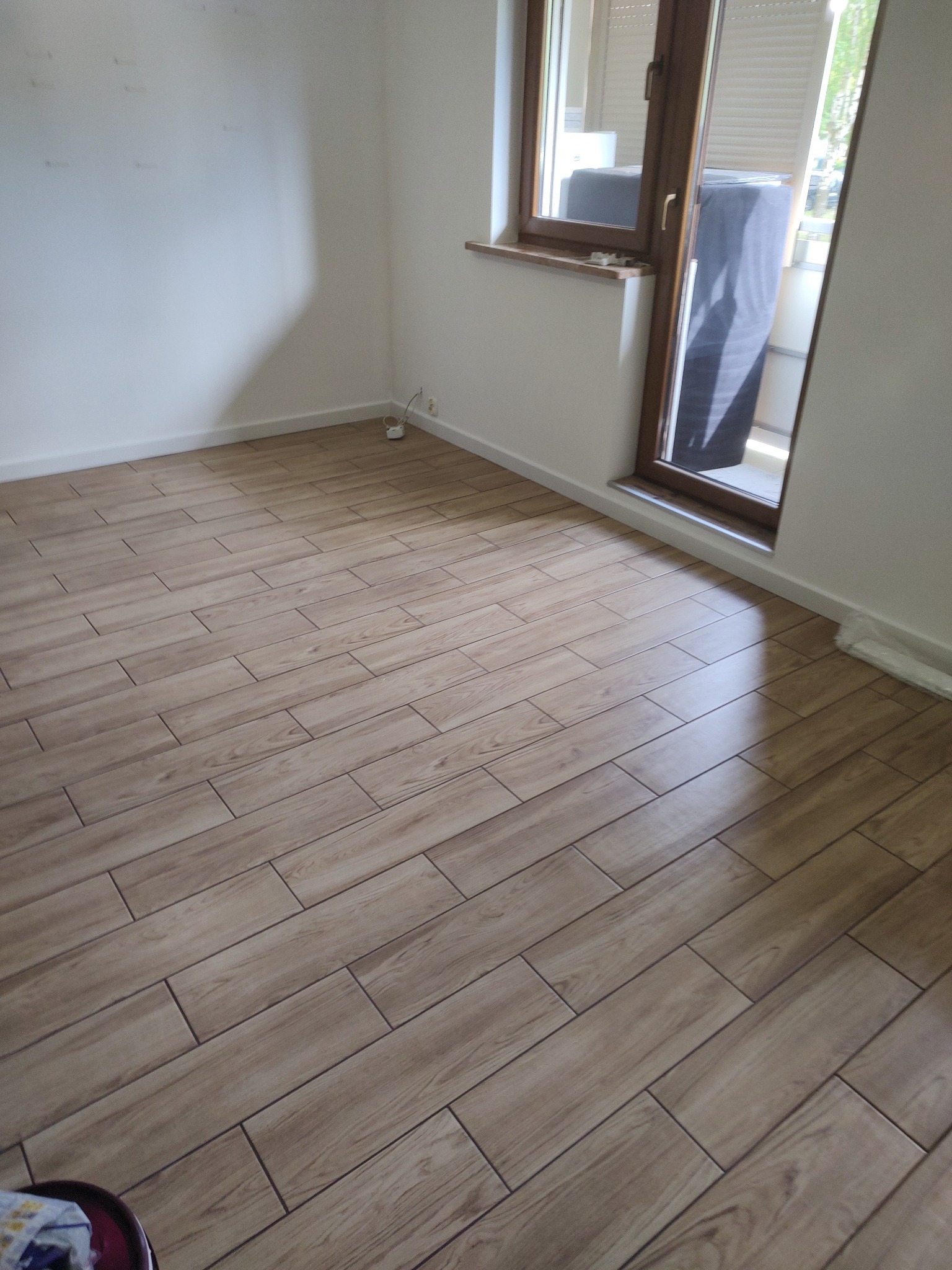 Ułożone panele podłogowe w jasnym odcieniu drewna w pustym pokoju z białymi ścianami i oknem balkonowym.