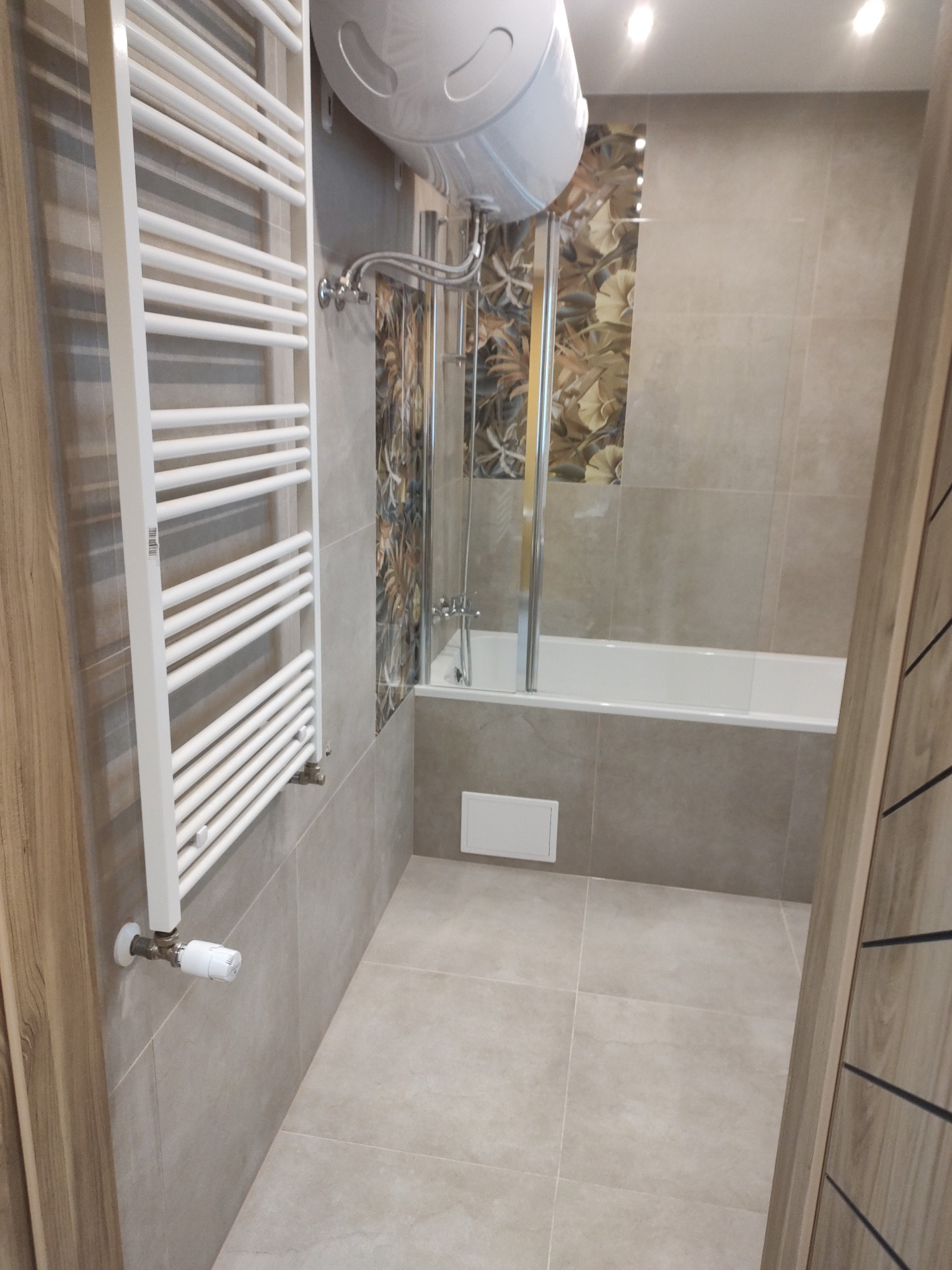 Wykończona łazienka z beżowymi płytkami, grzejnikiem drabinkowym, wanną z parawanem nawannowym oraz bojlerem elektrycznym nad wanną.