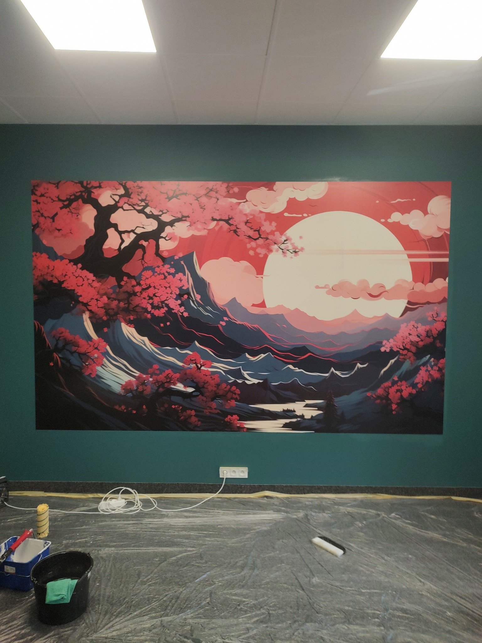 Ściana pomalowana na ciemnozielono z dużym, kolorowym obrazem w stylu japońskim przedstawiającym krajobraz z kwitnącymi drzewami, na podłodze folia ochronna i akcesoria malarskie, w tym wiadro...
