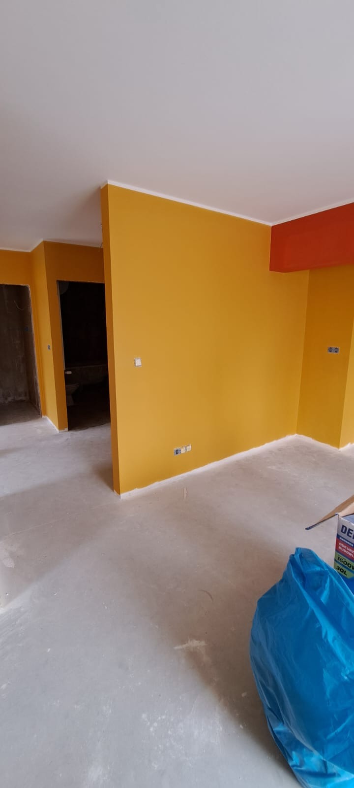 Świeżo pomalowane na żółto ściany pomieszczenia z fragmentem sufitu w kolorze białym i pomarańczowym, widoczne gniazdka elektryczne, surowa podłoga i niebieski worek z folii na podłodze.