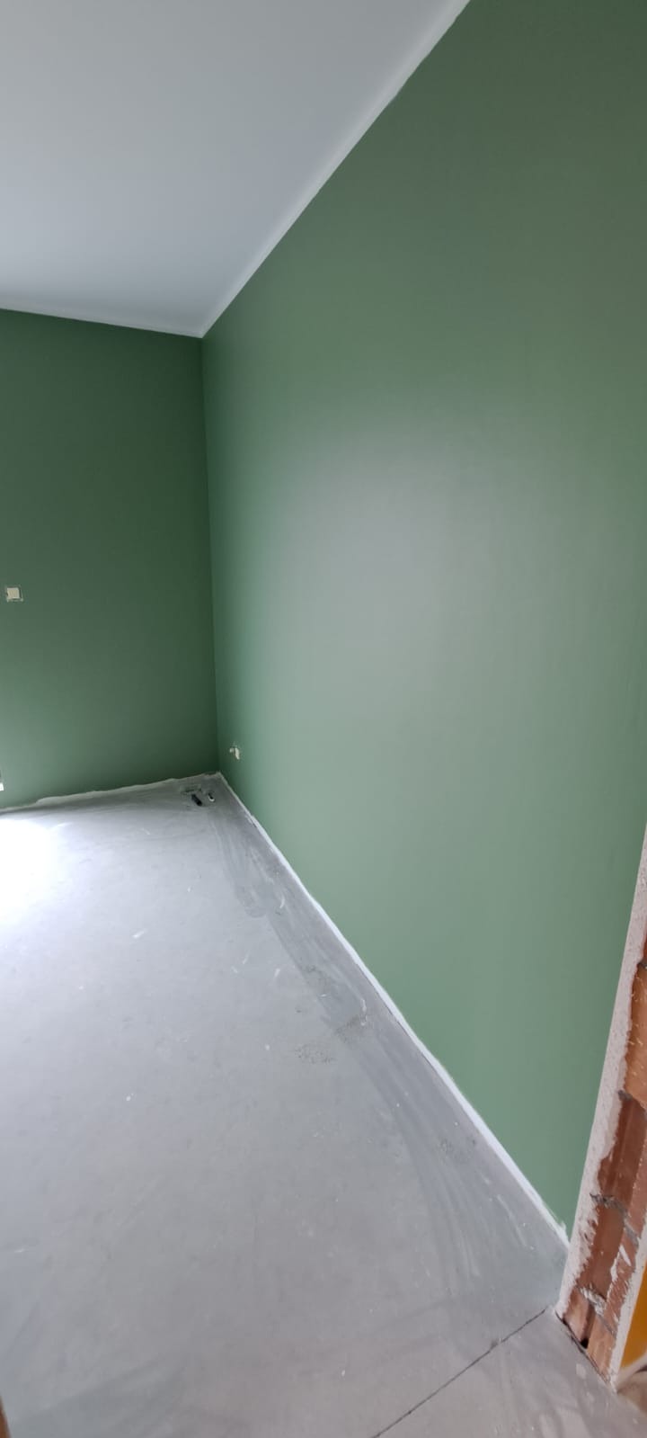 Pomalowane na zielono ściany w pustym pokoju z szarą podłogą przygotowaną do dalszych prac wykończeniowych, widoczne fragmenty cegieł przy futrynie.