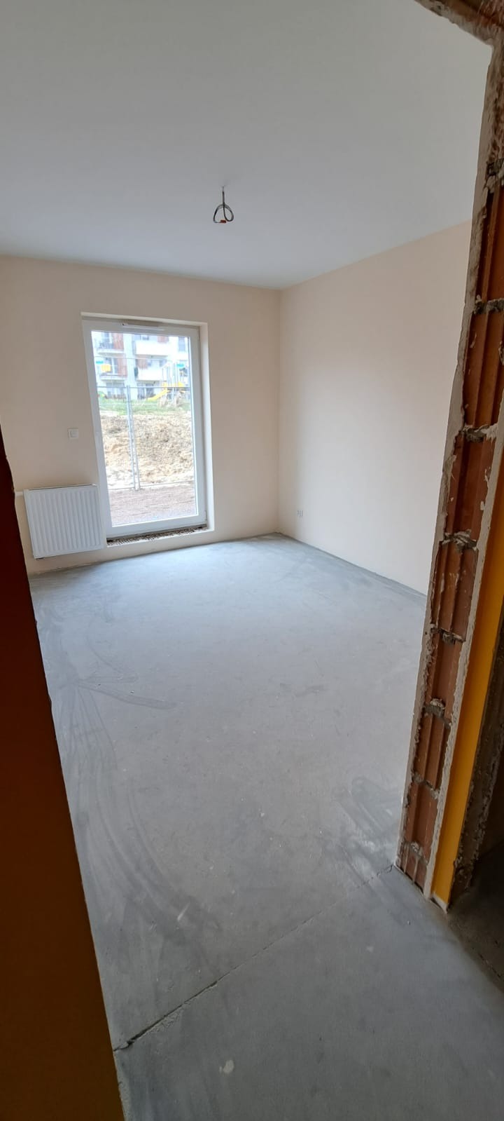 Pusty pokój po malowaniu na jasny beż, z surową wylewką na podłodze i widoczną instalacją elektryczną na suficie, okno z grzejnikiem, widok na plac budowy za oknem.