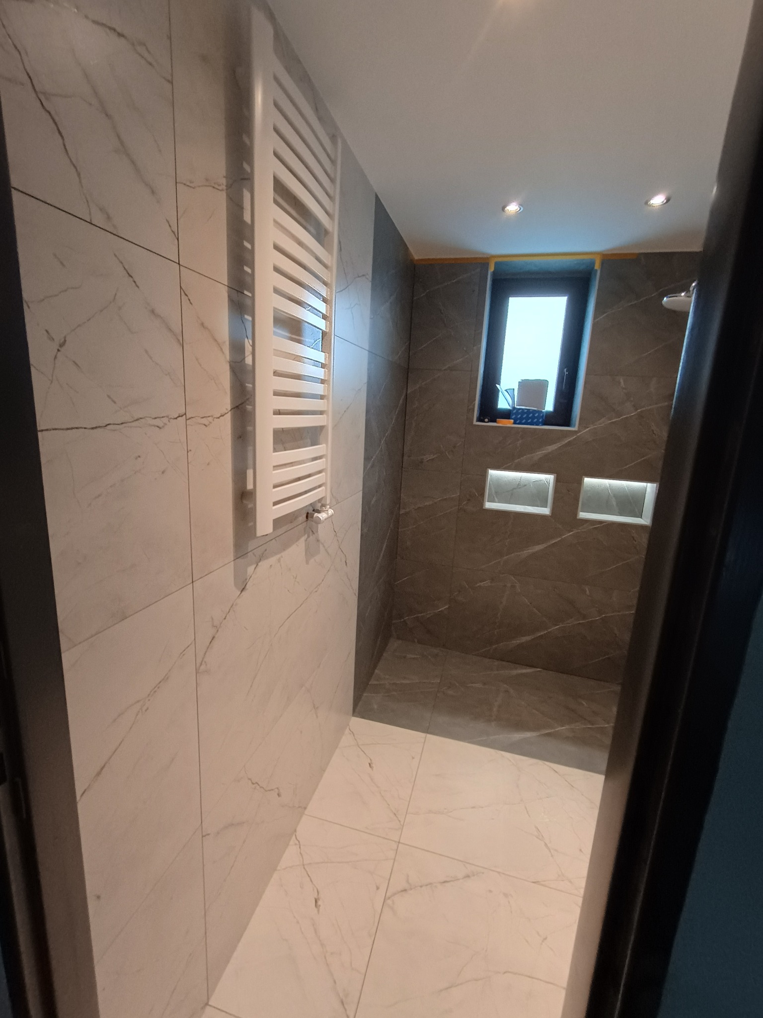 Wykończona łazienka z marmurowymi płytkami, grzejnikiem drabinkowym i oknem. Wnęki z oświetleniem. Widok zza futryny.