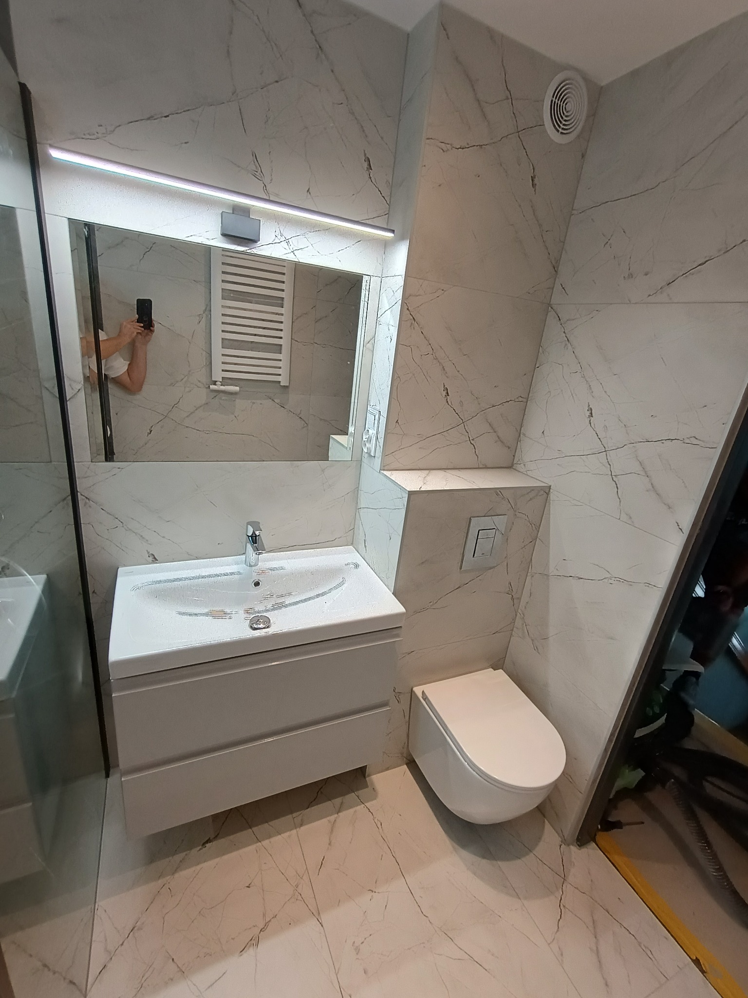 Wykończona łazienka z białymi płytkami imitującymi marmur, podwieszana toaleta, umywalka z szafką i lustro z oświetleniem LED. Widoczny fragment drzwi kabiny prysznicowej.