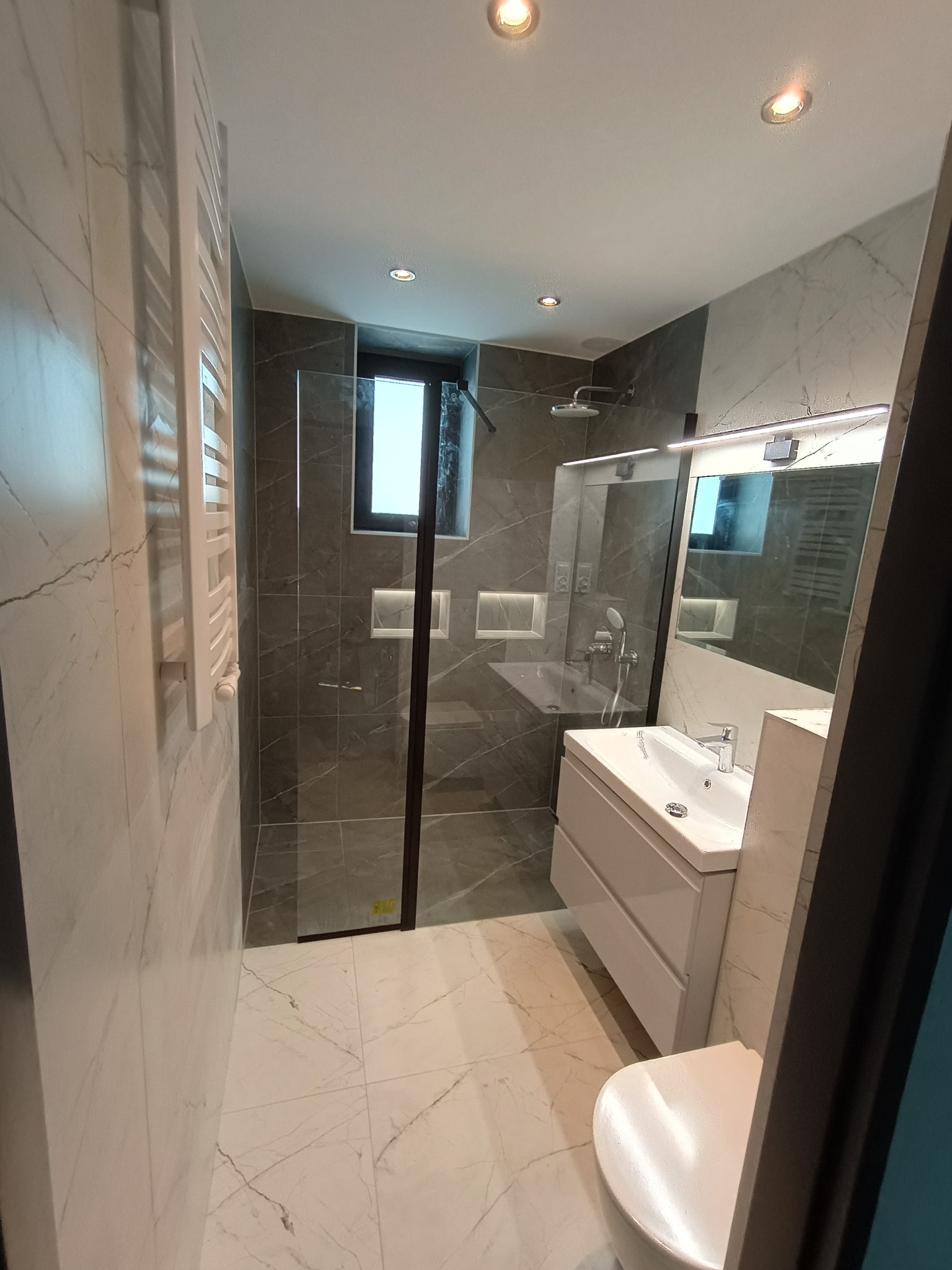 Nowoczesna łazienka w szaro-białej tonacji z kabiną prysznicową typu walk-in, umywalką z szafką i grzejnikiem drabinkowym. Marmurowe wykończenie ścian i podłóg.